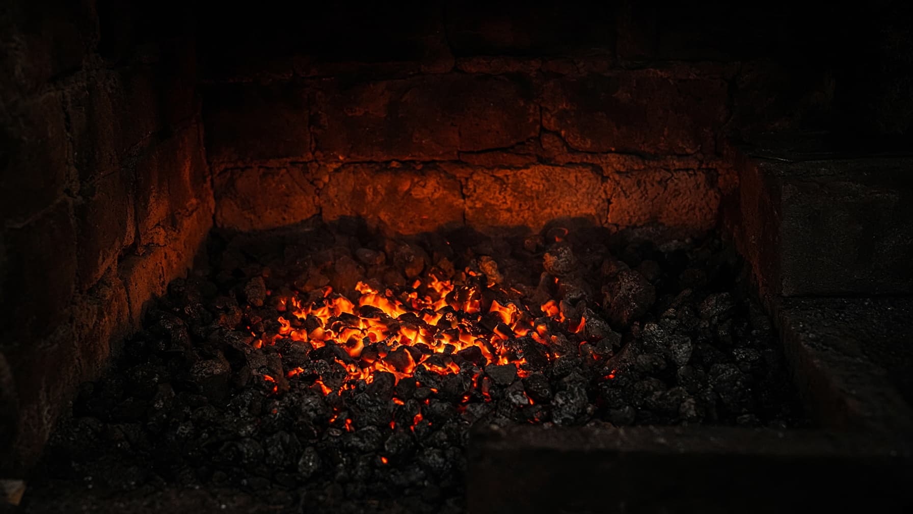 A blacksmith's forge hearth glowing in near darkness, orange embers among dark coals.